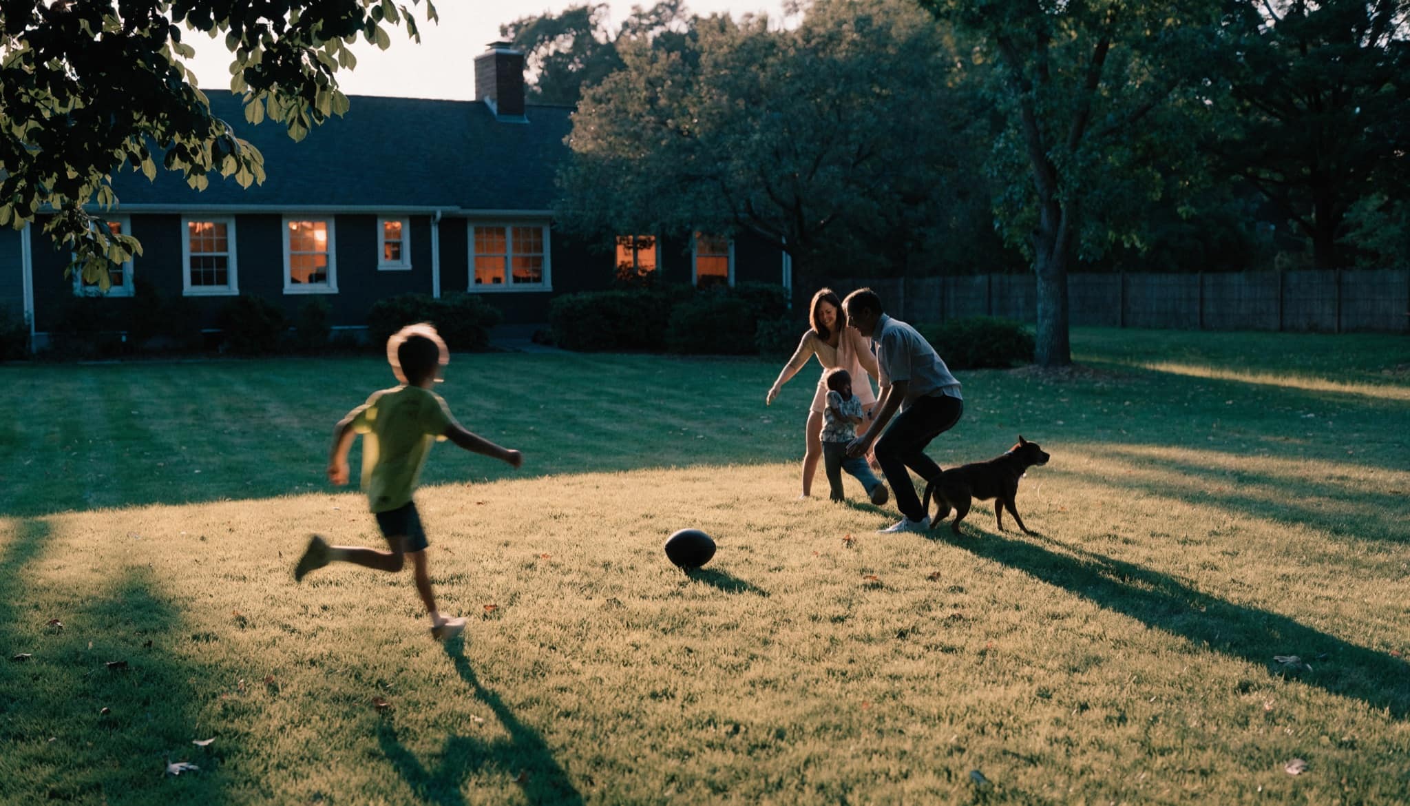 A family playing football together on the lawn behind their home at golden hour, the windows lit warmly behind them